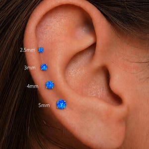 May include: Four round blue stud earrings arranged vertically on an earlobe, labeled with sizes 2.5mm, 3mm, 4mm, and 5mm. Each earring features a vibrant blue stone set in a simple metal prong setting, showcasing the size progression clearly.