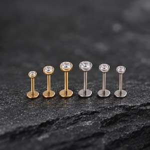 May include: A collection of six labret studs arranged in a row, featuring three gold-toned and three silver-toned metal posts. Each stud is topped with a single clear, round-cut gemstone set in a bezel, with a flat circular base at the bottom.