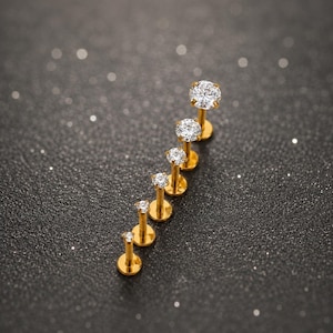 May include: A set of six gold-toned labret studs arranged in a vertical line, each featuring a single round clear gemstone set in a prong setting. The studs vary in size, with the largest at the top and the smallest at the bottom of the row.