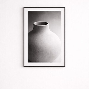 May include: Black and white photograph of a large, rounded vase with a narrow neck. The vase has a textured, matte finish and is set against a soft, gradient background. The artwork is framed in a simple black frame.