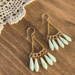 May include: A pair of gold-colored earrings with a chandelier design. Each earring features a series of small, light green teardrop-shaped beads suspended from a gold chain. The earrings are displayed on a wooden surface.