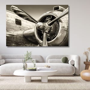 Könnte beinhalten: Ein Wandkunstdruck in Sepiatönen zeigt eine Nahaufnahme eines alten Flugzeugmotors und Propellers. Die metallische Oberfläche des Flugzeugs weist Nieten und Paneele auf, wobei die großen Propellerblätter von der zentralen Nabe ausgehen.