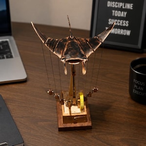 May include: A mechanical kinetic sculpture featuring a dark bronze-coloured manta ray suspended above a wooden base. The manta ray is connected by thin wires to a series of small metal components and a bright yellow gear mechanism on the base.