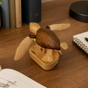 May include: A wooden sea turtle figurine featuring a dark brown carved shell and lighter tan flippers. The turtle is mounted on a light-colored oval wooden base with a small yellow accent visible near the connection point of the front flipper.
