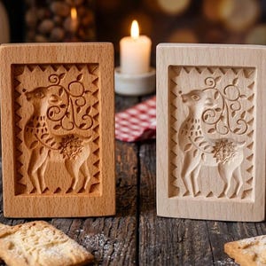 May include: Two rectangular wooden biscuit moulds featuring a carved deer design with intricate floral antlers and a star pattern on the body. The moulds are light brown and beige, standing upright with a serrated border around the central deer figure.