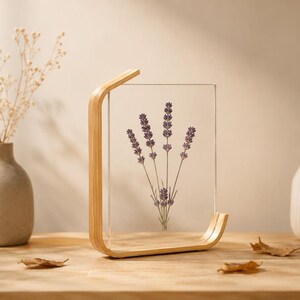 Puede incluir: Un marco rectangular de acrílico transparente sostenido por un soporte de madera clara curvada. Dentro del acrílico transparente, se exhibe una rama prensada de flores de lavanda moradas. El marco está colocado en posición vertical.