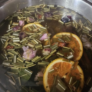 May include: A stainless steel pot filled with water and various ingredients. Sliced orange halves, pink flower buds, and green herbs float in the water. The water is a light brown color, and the pot is partially visible.