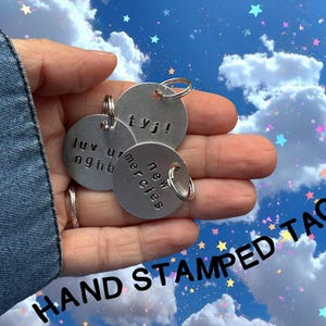 May include: Three silver, hand-stamped metal tags with various phrases, held in a hand. The tags are round and attached to silver rings. The background is a blue sky with clouds and stars, and the words "HAND STAMPED TAGS" are printed in black.