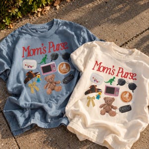 May include: A blue and a cream t-shirt, each featuring the red text Mom's Purse above a graphic collage of items including a green toy dinosaur, black sunglasses, a pink toy, a coffee cup, a brown teddy bear, a car key, and a hair clip.