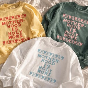 May include: Three crewneck sweatshirts in yellow, sage green, and white. Each features the text MOTHERHOOD IS HOLY WORK in a bold font, framed by horizontal bands of heart and snowflake patterns in red, light pink, and light blue respectively.