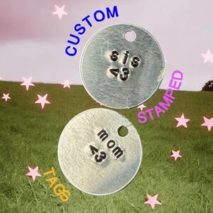 May include: Two silver custom stamped tags with the words "sis <3" and "mom <3" stamped on them. The tags are round with a hole at the top. The word "CUSTOM" is written in blue above the top tag, "STAMPED" in pink, and "TAGS" in yellow.