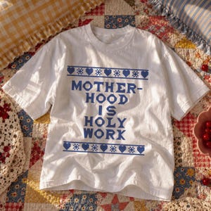 May include: A white short-sleeved t-shirt featuring blue text that reads MOTHERHOOD IS HOLY WORK. The text is centred and framed by two horizontal rows of blue geometric star and heart symbols, creating a decorative border above and below.