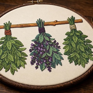 Peut inclure: Un tambour à broder contenant un tissu blanc cassé avec un motif brodé de trois bouquets d'herbes suspendus à une branche en bois horizontale. Les bouquets présentent des feuilles vertes, des fleurs violettes et sont liés par du fil orange, violet et jaune.