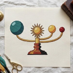 Peut inclure: Une broderie au point de croix sur toile Aida blanche représentant un modèle d'orrery céleste. Le motif présente un soleil central avec des rayons, flanqué de trois sphères en orbite bleu sarcelle, jaune et rouge, reliées par des bras dorés.