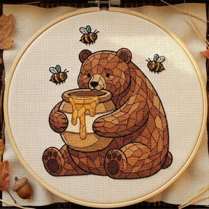 Peut inclure: Une broderie au point de croix dans un tambour en bois circulaire représentant un ours brun au motif géométrique tenant un pot de miel avec du miel doré qui coule, entouré de trois petites abeilles sur un tissu de couleur crème.