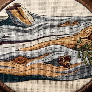 Puede incluir: Un bordado de punto de cruz en un bastidor de madera circular que muestra un tronco desgastado con corteza texturizada en tonos bronceado, gris y verde oscuro, acentuado por pequeñas bayas rojas y una rama de agujas de pino verdes.