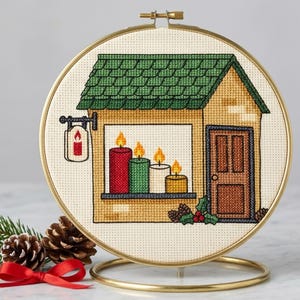 Peut inclure: Une broderie au point de croix dans un tambour en bois circulaire représentant une maison avec un toit vert, une porte marron et une fenêtre montrant quatre bougies allumées rouges, vertes, blanches et jaunes avec un petit panneau.
