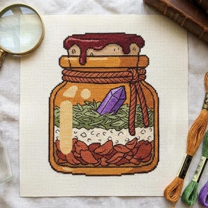 Peut inclure: Un motif de point de croix sur tissu blanc représentant un bocal en verre avec un sceau en cire rouge foncé. À l'intérieur se trouvent des couches de pétales rouges, de grains blancs et d'herbes vertes, avec un cristal violet posé au-dessus.