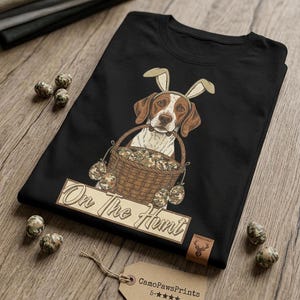 Peut inclure: T-shirt noir avec un chien portant des oreilles de lapin et tenant un panier d'œufs de Pâques camouflage. Le texte "On The Hunt" est en dessous du panier. Une étiquette avec "CamoPawsPrints" et des étoiles est attachée.
