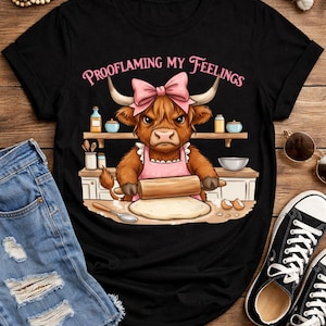 May include: Black t-shirt with a graphic of a Highland cow wearing a pink bow and apron, rolling dough. The text "Prooflaming My Feelings" is arched above the cow. The image is on a wooden surface with jeans and sneakers.