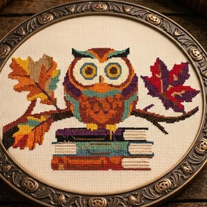 May include: A cross-stitch embroidery featuring a colorful owl perched on a branch with autumn leaves, resting atop a stack of three books. The design is framed in a circular, dark metallic hoop decorated with small embossed pumpkin motifs.
