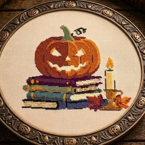 May include: A cross-stitch embroidery featuring a carved orange jack-o'-lantern pumpkin resting on a stack of four books, accompanied by a lit candle in a gold holder and autumn leaves, all framed in a round, dark metal frame with skull motifs.