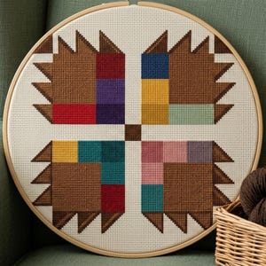 May include: A circular wooden embroidery hoop containing a cross-stitch pattern on white fabric. The design features four symmetrical brown geometric shapes arranged around a central brown square, accented with blocks of red, purple, yellow, teal, and pink.