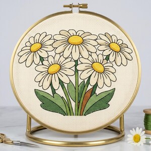 Peut inclure: Un tambour à broder circulaire en métal doré maintient un morceau de tissu Aida crème. Le tissu présente un motif au point de croix de cinq marguerites blanches aux centres jaune vif et aux feuilles vertes, posé sur un support doré.