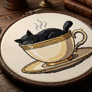 Peut inclure: Une broderie au point de croix dans un tambour en bois circulaire représentant un chat noir recroquevillé dans une tasse à thé crème avec une bordure dorée. La tasse repose sur une soucoupe assortie et de la vapeur s'échappe du chat.
