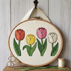 Peut inclure: Un tambour à broder circulaire en bois contenant un motif au point de croix de quatre tulipes orange, jaunes, roses et blanches avec des tiges et des feuilles vertes sur tissu crème. Un ruban crème est fixé au sommet et des ciseaux dorés sont posés.