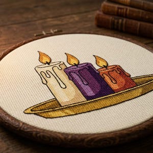 Peut inclure: Une broderie au point de croix dans un tambour en bois circulaire représentant trois bougies allumées de couleur blanche, violette et orange, posées sur un plateau doré. Les bougies présentent des coulures de cire et des flammes jaunes.