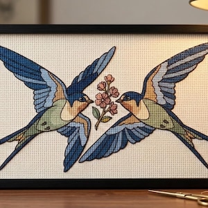 Peut inclure: Une broderie au point de croix dans un cadre noir représentant deux hirondelles aux ailes bleues, vertes et fauves se faisant face. Entre les oiseaux se trouve une petite branche avec des fleurs roses sur un tissu Aida crème.