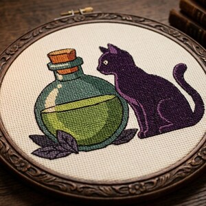 Peut inclure: Un tambour à broder circulaire contenant un motif au point de croix sur tissu blanc cassé. Le motif représente une bouteille en verre ronde remplie de liquide vert, un bouchon en liège et un chat violet assis à côté, orné de petites feuilles violettes.