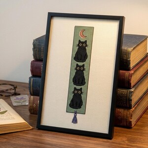 Peut inclure: Un marque-page en point de croix encadré représentant trois chats noirs empilés sur un fond vert sauge avec un petit croissant de lune orange en haut. Le marque-page possède un pompon bleu clair en bas et est dans un cadre noir fin.