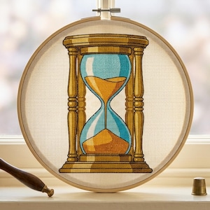 Peut inclure: Un projet de broderie au point de croix présenté dans un tambour en bois circulaire, représentant un sablier détaillé avec un cadre doré et des sections en verre bleu vif contenant du sable jaune doré s'écoulant entre les deux chambres.