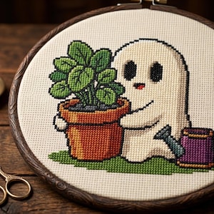 Peut inclure: Une broderie au point de croix dans un tambour en bois circulaire représentant un fantôme blanc tenant un pot couleur terre cuite avec une plante verte. Un petit arrosoir violet est posé sur l'herbe verte brodée à côté du fantôme.