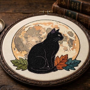 Peut inclure: Un tambour à broder circulaire présentant un motif au point de croix d'un chat noir assis devant une grande pleine lune texturée beige et ocre. Le chat est entouré de trois feuilles d'automne stylisées aux tons vert, orange et bleu canard.