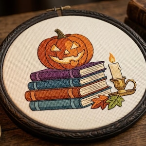 May include: A circular embroidery hoop containing a cross-stitch design of a carved orange jack-o'-lantern resting on a stack of three books in purple, blue, and orange. Beside the books is a lit candle in a gold holder and two fallen leaves.