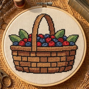 May include: A cross-stitch embroidery piece in a circular wooden hoop featuring a brown woven basket filled with red and blue berries and green leaves. The design is stitched onto a light beige fabric with a visible grid pattern throughout.