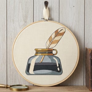 Peut inclure: Un tambour à broder circulaire en bois contenant un motif au point de croix représentant un encrier en verre avec un bord doré et une plume rayée marron et blanche insérée sur le dessus, sur un fond en tissu de couleur crème.