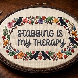 May include: An oval embroidery hoop containing cream-colored fabric with the dark gray stitched text STABBING IS MY THERAPY. The text is surrounded by a colorful embroidered wreath featuring black crows, autumn leaves, and various small flowers.