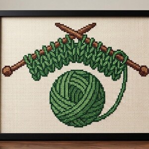 Peut inclure: Une broderie au point de croix encadrée en noir, représentant une pelote de laine verte et une rangée de mailles tricotées vertes sur deux aiguilles à tricoter marron. Le motif est centré sur une toile Aida crème avec une grille visible.
