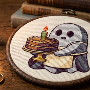 Peut inclure: Un tambour à broder circulaire en bois contenant un motif au point de croix représentant un fantôme gris clair portant un tablier crème. Le fantôme tient un gâteau marron sur un support doré, surmonté d'une bougie allumée à flamme orange.