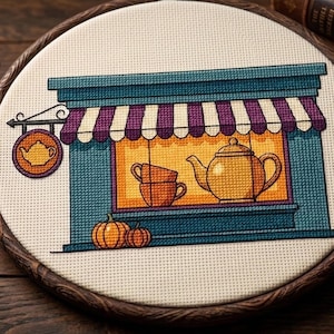 Peut inclure: Une broderie au point de croix dans un tambour en bois circulaire représentant une devanture de magasin avec une façade bleu sarcelle, un auvent rayé violet et blanc, et une vitrine orange lumineuse avec une théière et deux tasses.