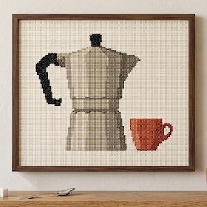 Peut inclure: Une broderie au point de croix encadrée représentant une cafetière italienne beige avec une anse noire et une petite tasse à café orange. Le motif est réalisé dans un style pixélisé sur un tissu crème, dans un cadre en bois brun foncé.