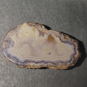 May include: A polished agate stone with a natural, irregular shape. The stone features concentric bands of white, lavender, and brown hues, with a rough, textured edge. The agate's surface is smooth and glossy.