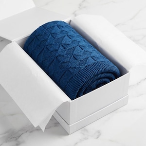 May include: A rolled navy blue knitted blanket with a geometric design, presented in a white gift box. The blanket's texture is visible, and the box is lined with white tissue paper. The presentation suggests a high-quality textile item, ideal for gifting.