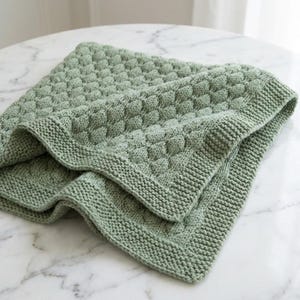 Può includere: Una coperta lavorata a maglia verde salvia con un motivo quadrato in rilievo strutturato. La coperta ha un bordo a coste ed è piegata, mostrando la dettagliata lavorazione a punto. Il colore morbido e tenue e la consistenza accogliente suggeriscono un articolo fatto a mano.