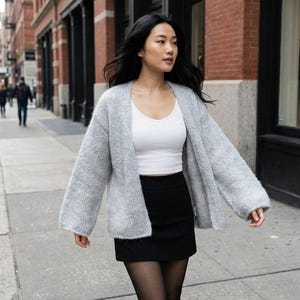 May include: A woman wearing a light gray, open-front cardigan, a white tank top, and a black skirt. She is walking on a city sidewalk. The cardigan has long sleeves and a loose fit. The background includes brick buildings and other pedestrians.