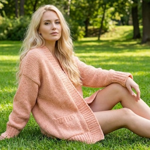 Op de afbeelding: Een licht perzikkleurig vest met open voorkant en twee voorzakken. Het vest is gemaakt van een dik gebreid materiaal. De persoon zit op groen gras in een park.